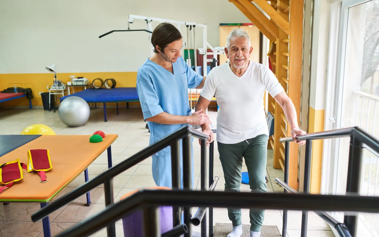 Physical therapist supporting an older man as he practices climbing steps with handrails
